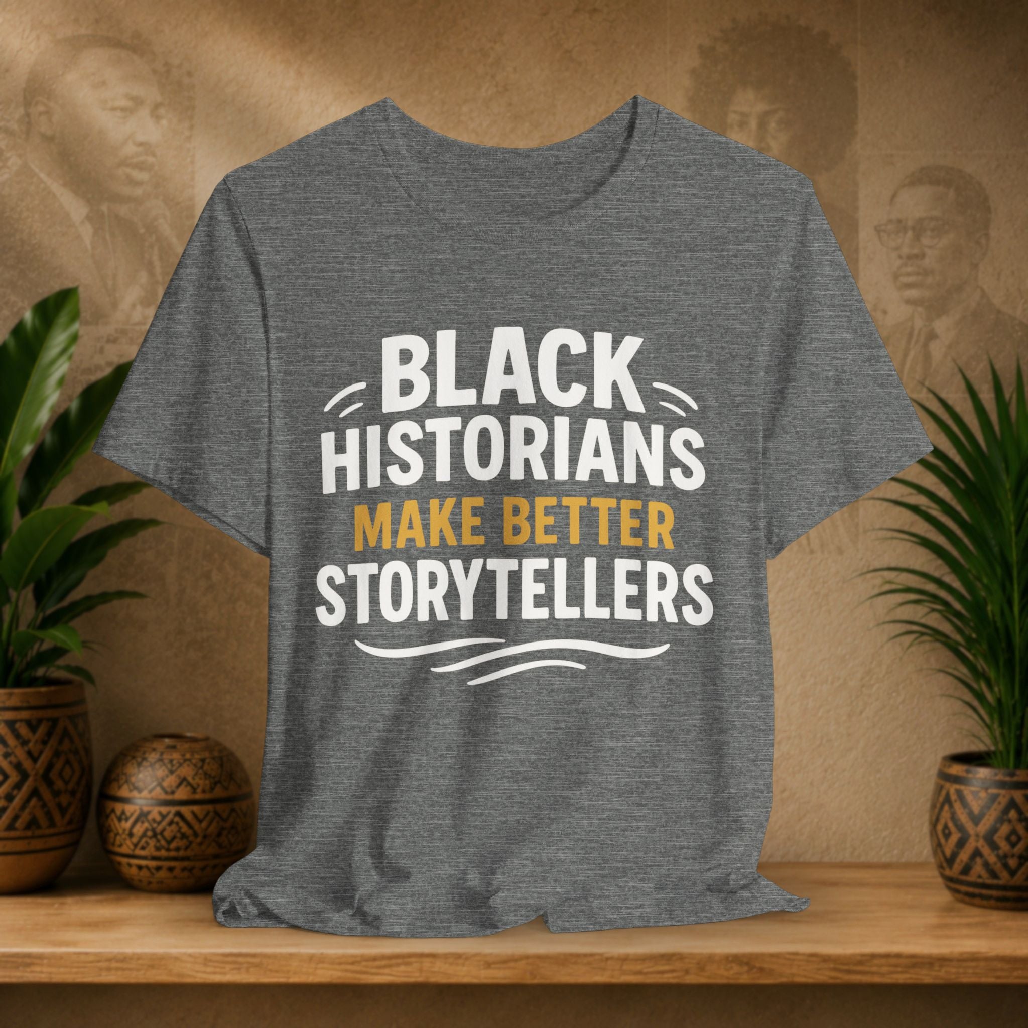 Gray t-shirt with 'Black Historians Make Better Storytellers' text on a wooden surface with plants and decorative items.