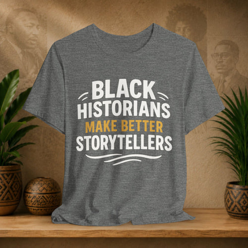 Gray t-shirt with 'Black Historians Make Better Storytellers' text on a wooden surface with plants and decorative items.