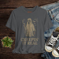 Gray t-shirt with ghost graphic and 'Creepin' It Real' text on a wooden surface with jeans and sneakers.