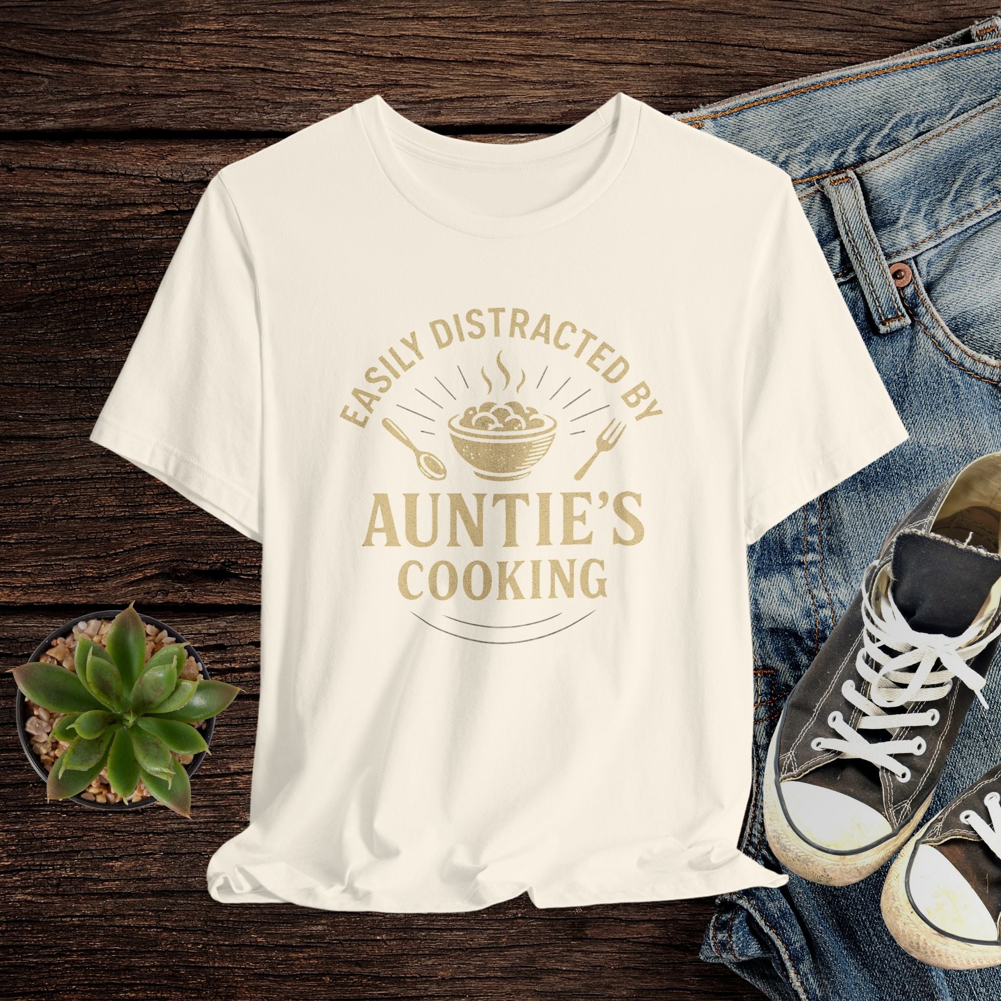 Beige t-shirt with 'Easily Distracted by Auntie's Cooking' text on a wooden surface with jeans and sneakers.