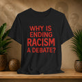 Black t-shirt with red text 'Why is ending racism a debate?' on a wooden surface with plants and a textured wall in the background.