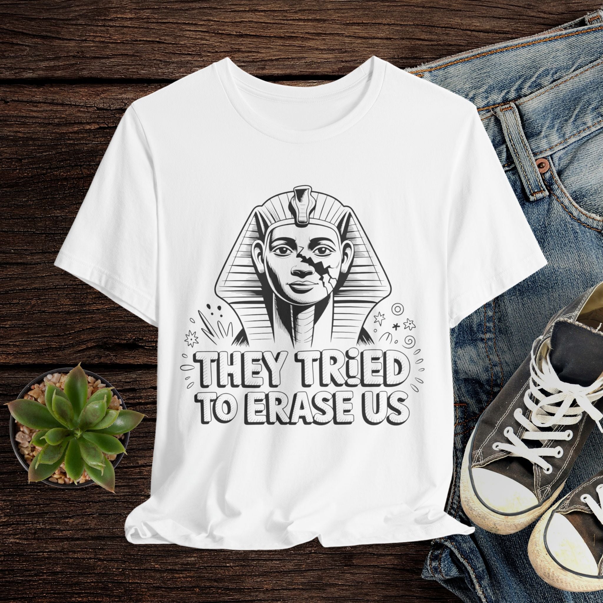 White t-shirt with Pharaoh graphic and text 'They tried to erase us' on a wooden surface with jeans and sneakers.