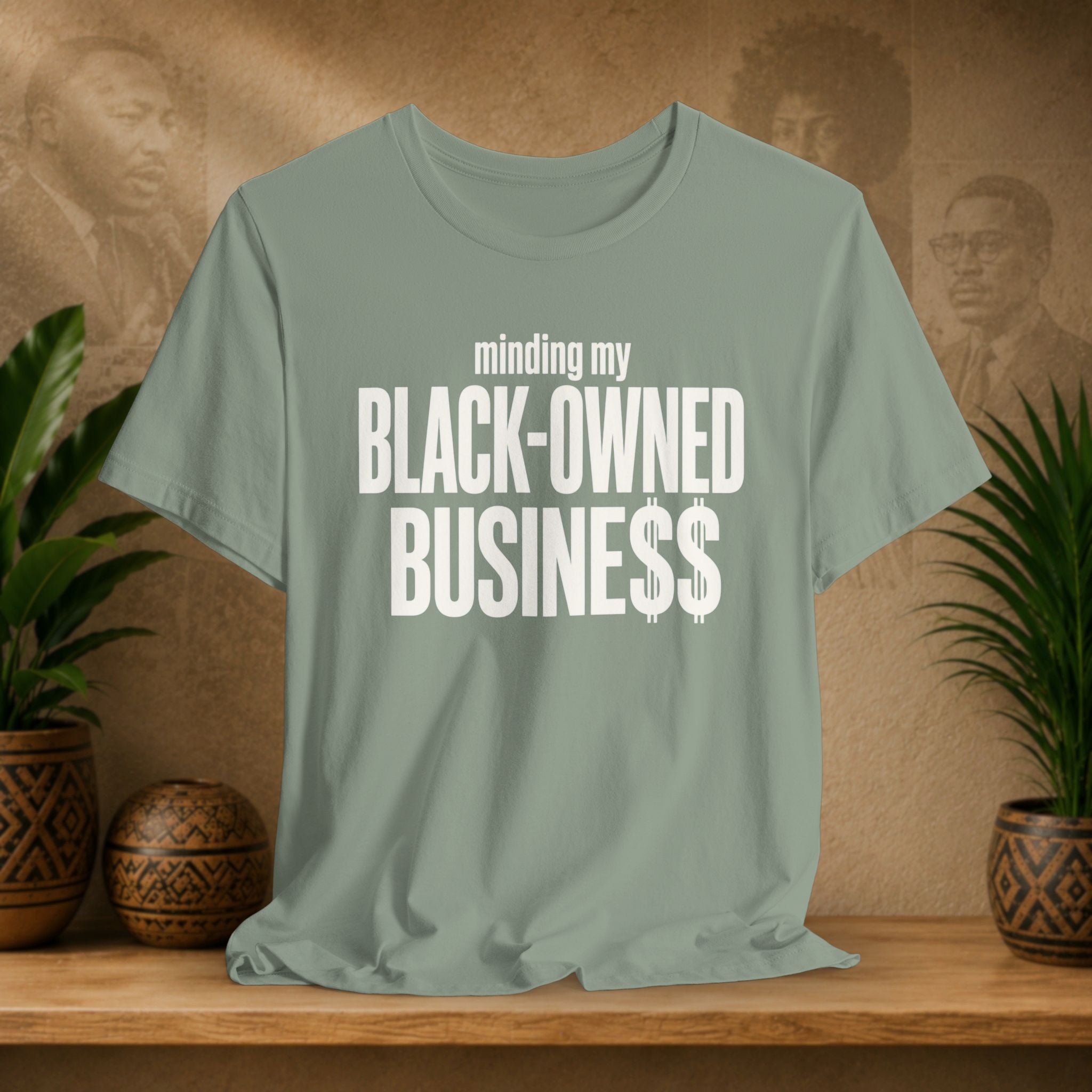 A sage green t-shirt with white text reading 'minding my BLACK-OWNED BUSINESS$$' displayed on a wooden shelf with potted plants and a textured background.