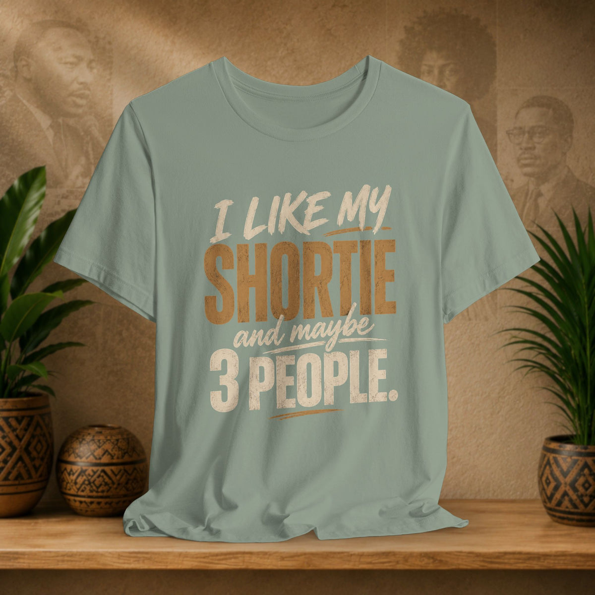 A sage graphic t-shirt displayed on a wooden shelf. The shirt features a humorous slogan printed in white and tan lettering: 'I LIKE MY SHORTIE and maybe 3 PEOPLE.