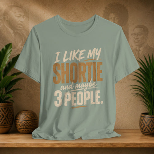A sage graphic t-shirt displayed on a wooden shelf. The shirt features a humorous slogan printed in white and tan lettering: 'I LIKE MY SHORTIE and maybe 3 PEOPLE.