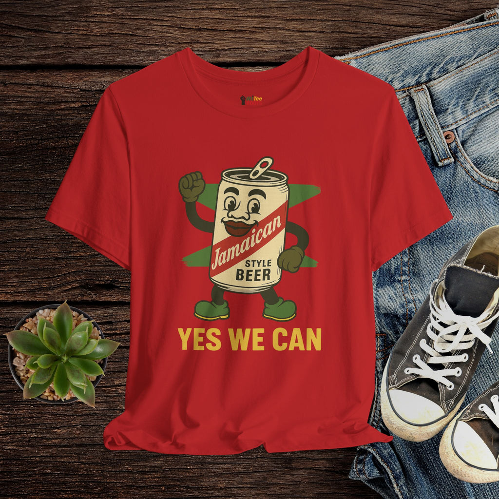 Red t-shirt with a cartoon beer can character and 'Yes We Can' text on a wooden surface with jeans and shoes.