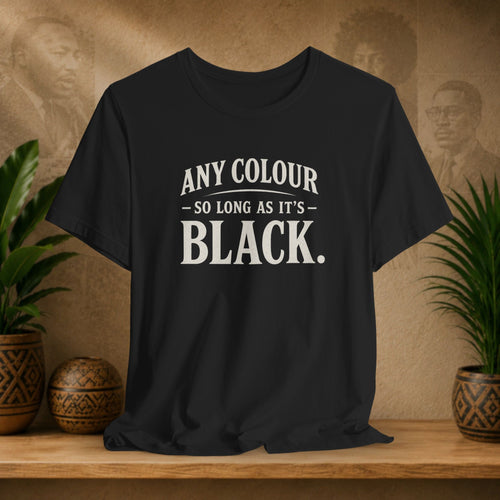 Black t-shirt with 'Any colour so long as it's black' text on a wooden surface with plants and a textured wall background.