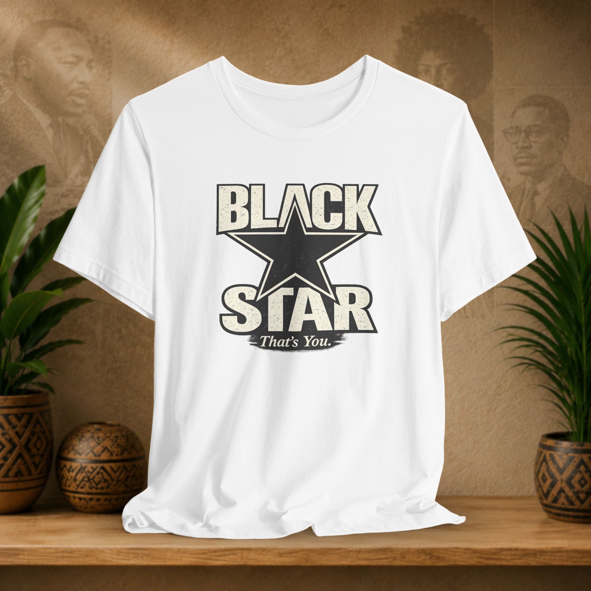 White t-shirt with 'Black Star That's You' design on a wooden surface with plants and a textured wall background.