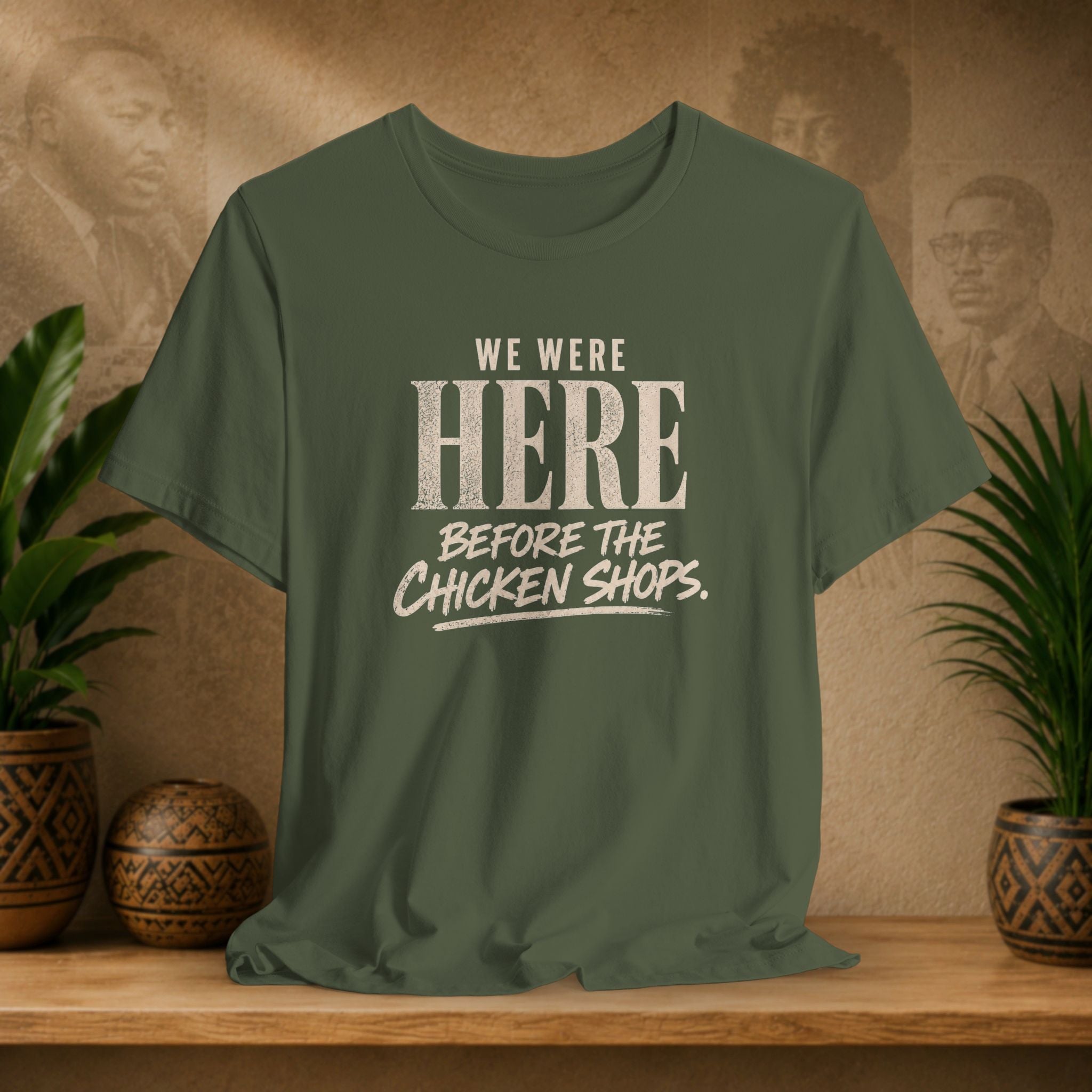 Green t-shirt with text 'WE WERE HERE BEFORE THE CHICKEN SHOPS' on a wooden surface with plants and a textured wall background.