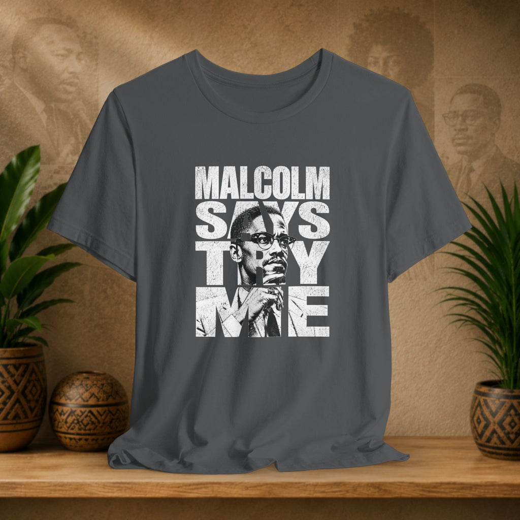 Gray t-shirt with 'Malcolm Says Thy Name' graphic on a wooden surface with plants and a textured wall in the background