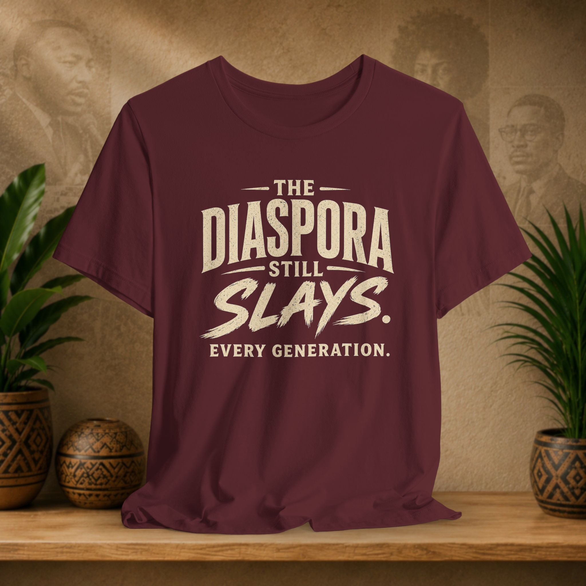 Maroon t-shirt with 'The Diaspora Still Slays Every Generation' text on a wooden surface with decorative plants and background.