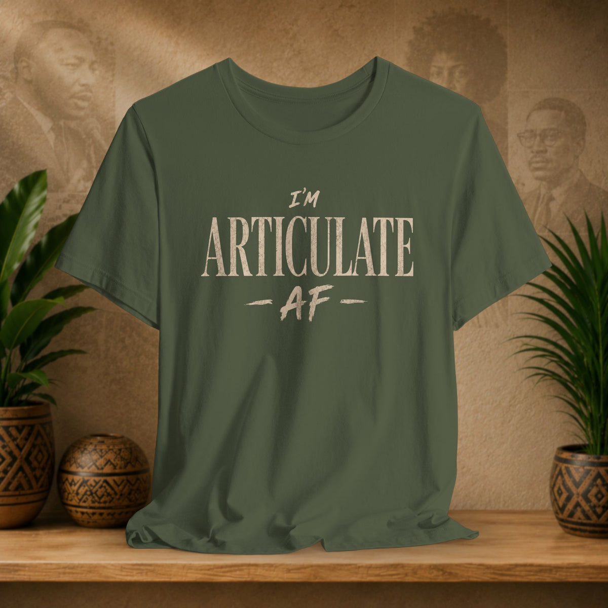 An olive green short-sleeve t-shirt with the text 'I'M ARTICULATE AF' printed in a distressed cream font, displayed on a wooden shelf with potted plants and a textured background.