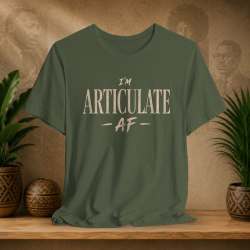 An olive green short-sleeve t-shirt with the text 'I'M ARTICULATE AF' printed in a distressed cream font, displayed on a wooden shelf with potted plants and a textured background.