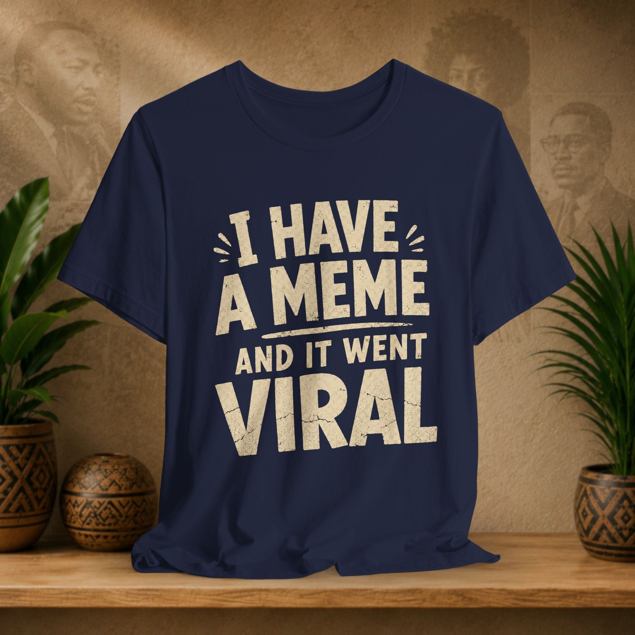 Navy blue t-shirt with white text 'I HAVE A MEME AND IT WENT VIRAL' on a wooden surface with plants and a textured wall background.