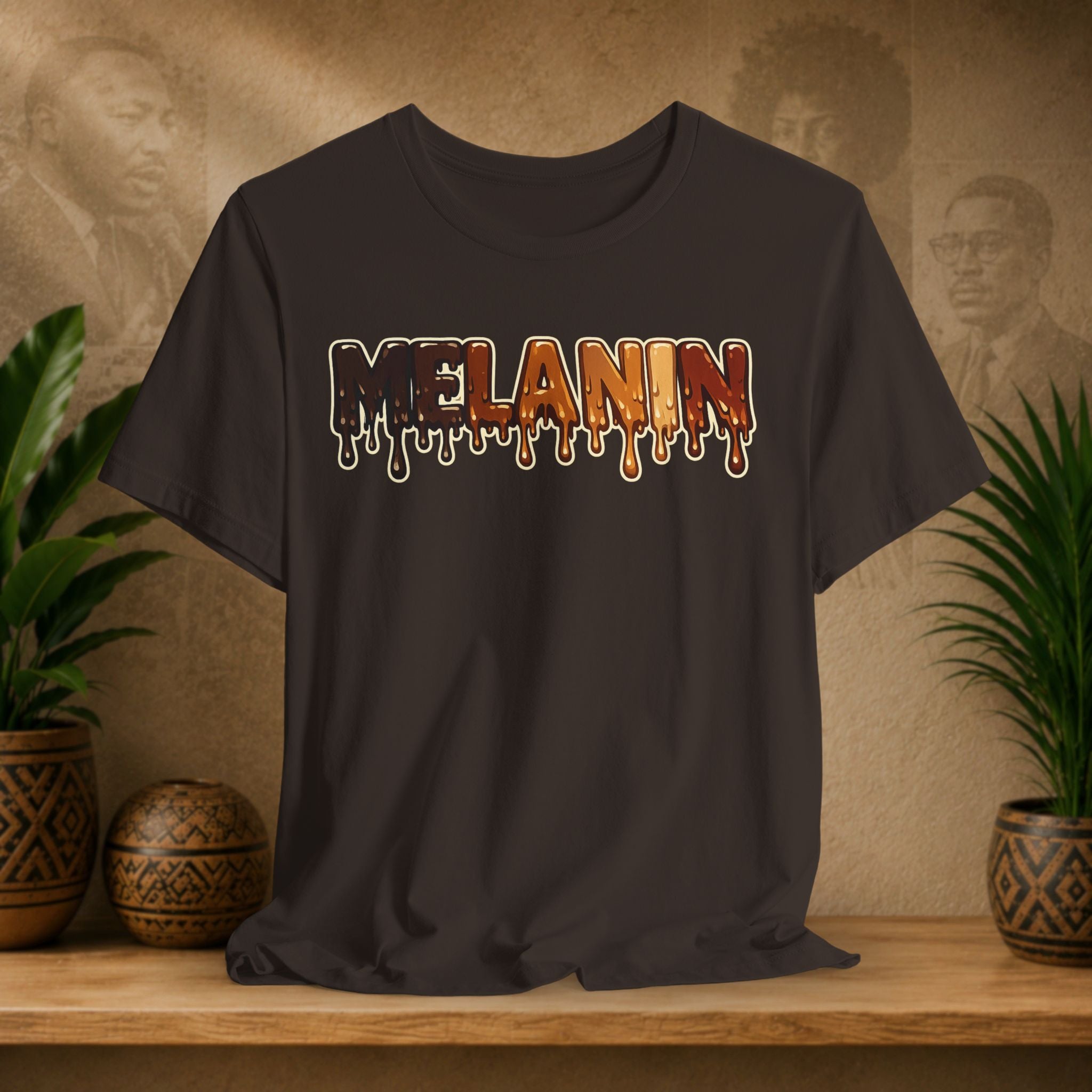 A brown t-shirt with a dripping 'MELANIN' graphic in orange, brown, and cream tones, displayed on a wooden shelf with potted plants against a textured beige background.