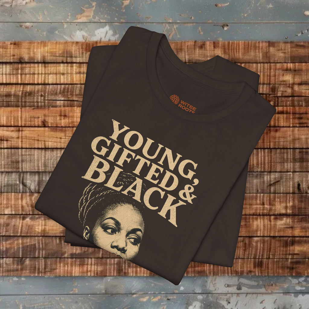 A folded brown graphic T-shirt with the words 'YOUNG, GIFTED & BLACK' in large beige lettering and a portrait graphic, displayed on a rustic wooden plank surface.