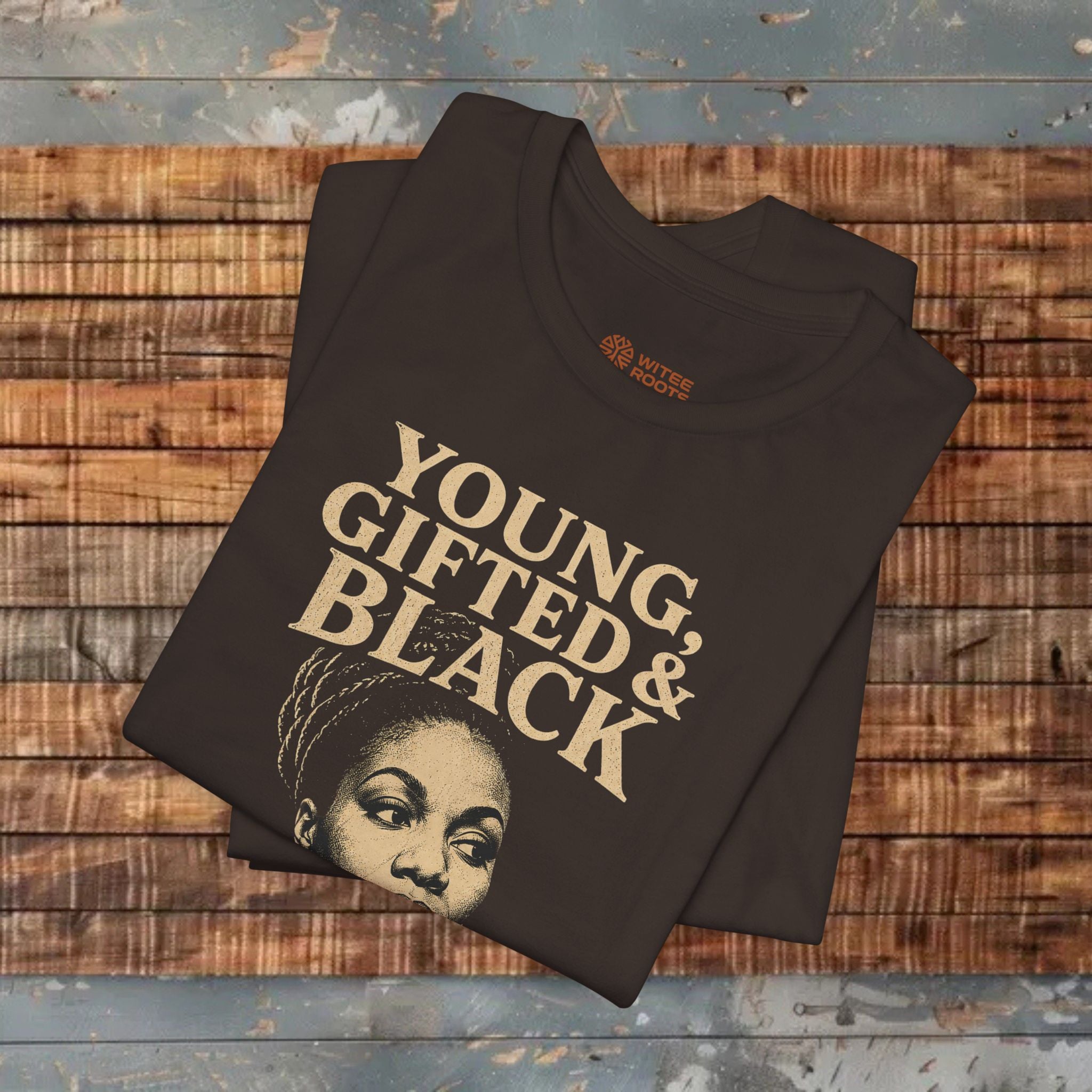 A folded brown graphic T-shirt with the words 'YOUNG, GIFTED & BLACK' in large beige lettering and a portrait graphic, displayed on a rustic wooden plank surface.