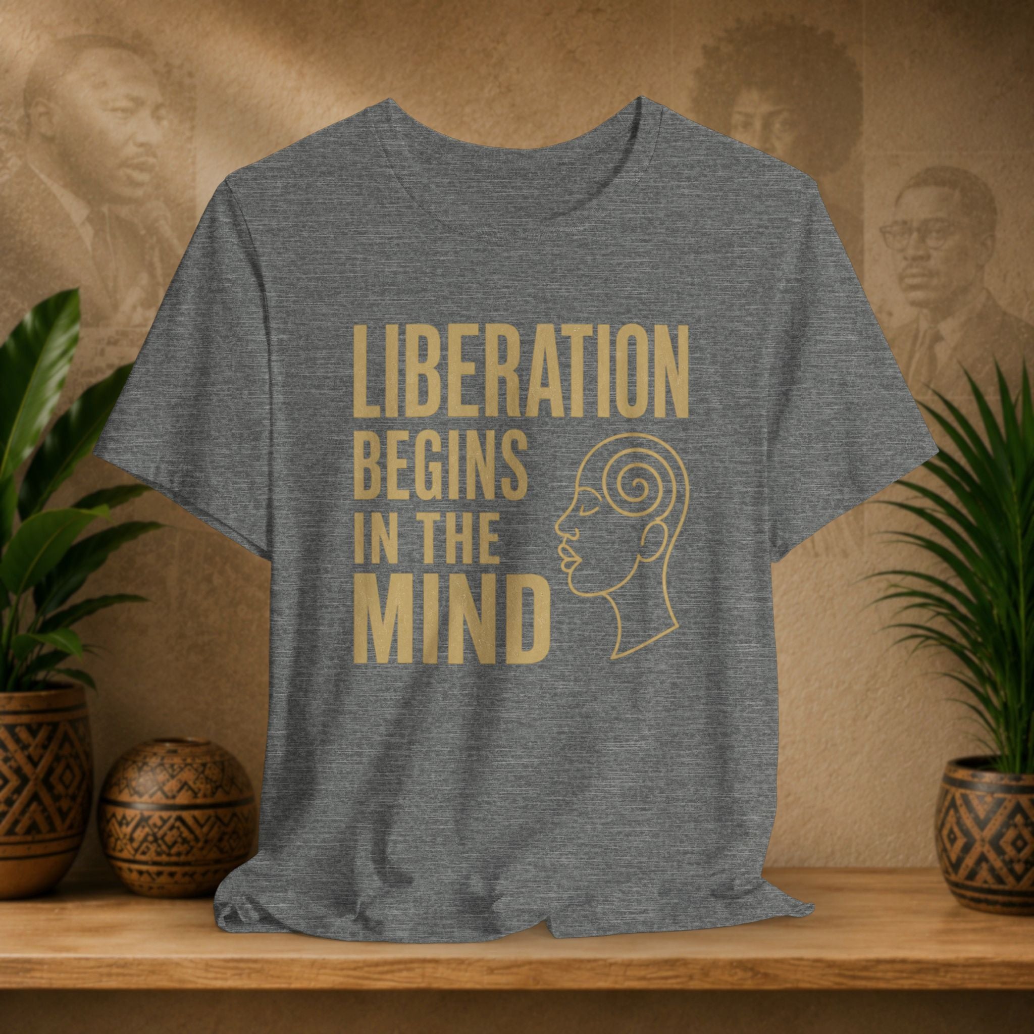 Gray t-shirt with 'Liberation Begins in the Mind' text and graphic on a wooden surface with plants and decorative items.