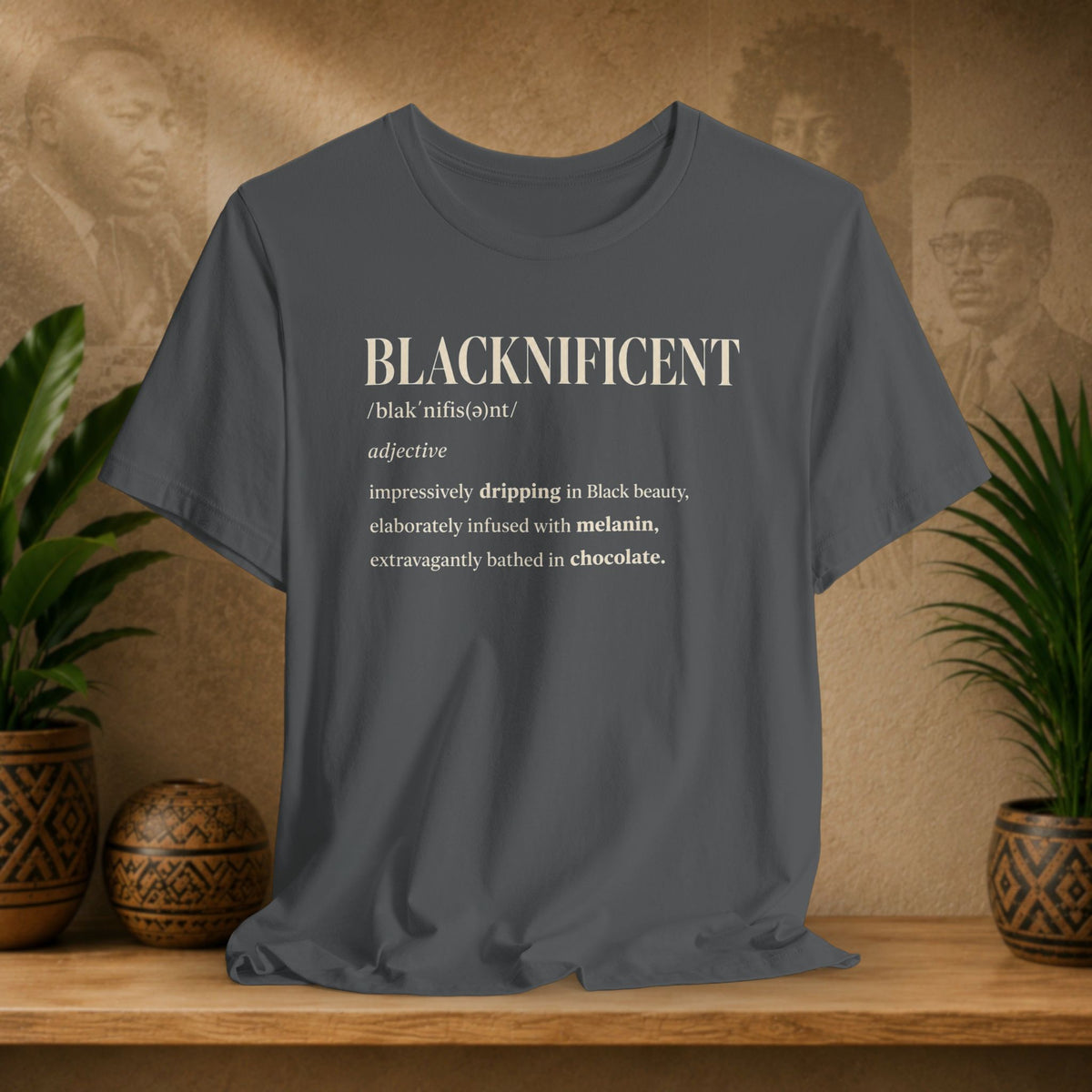 A dark gray graphic t-shirt displayed on a wooden shelf, flanked by potted green plants against a textured beige background with sepia portraits. The shirt features a cream-colored typographic print on the front reading 'BLACKNIFICENT' with a dictionary definition.