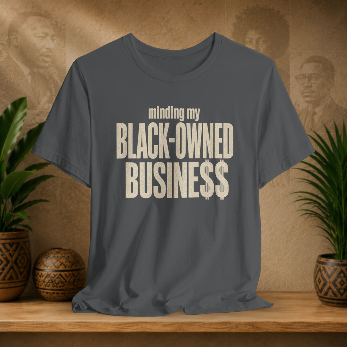 A dark gray t-shirt with the text 'minding my BLACK-OWNED BUSINESS$$' printed in distressed cream lettering, displayed on a wooden shelf with potted plants against a textured beige background.