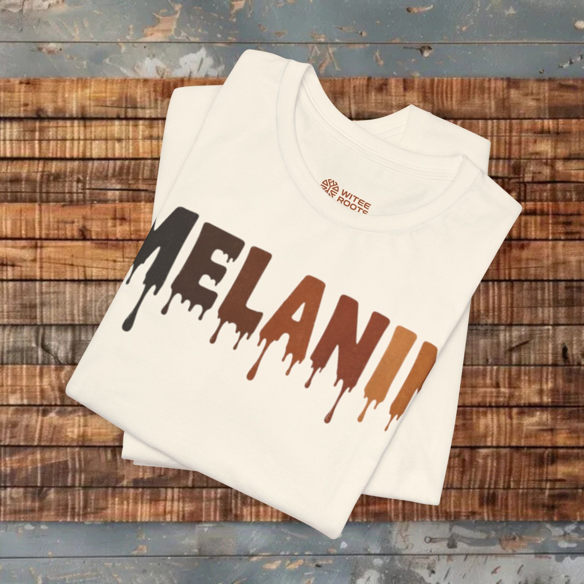 A folded beige t-shirt with the word 'MELANIE' printed in a dripping paint style transitioning from black to brown to orange. The shirt is displayed on a rustic wooden plank surface.