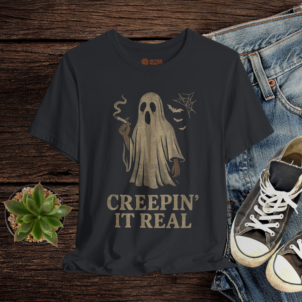 Black t-shirt with ghost graphic and 'Creepin' It Real' text on a wooden surface with jeans and sneakers.