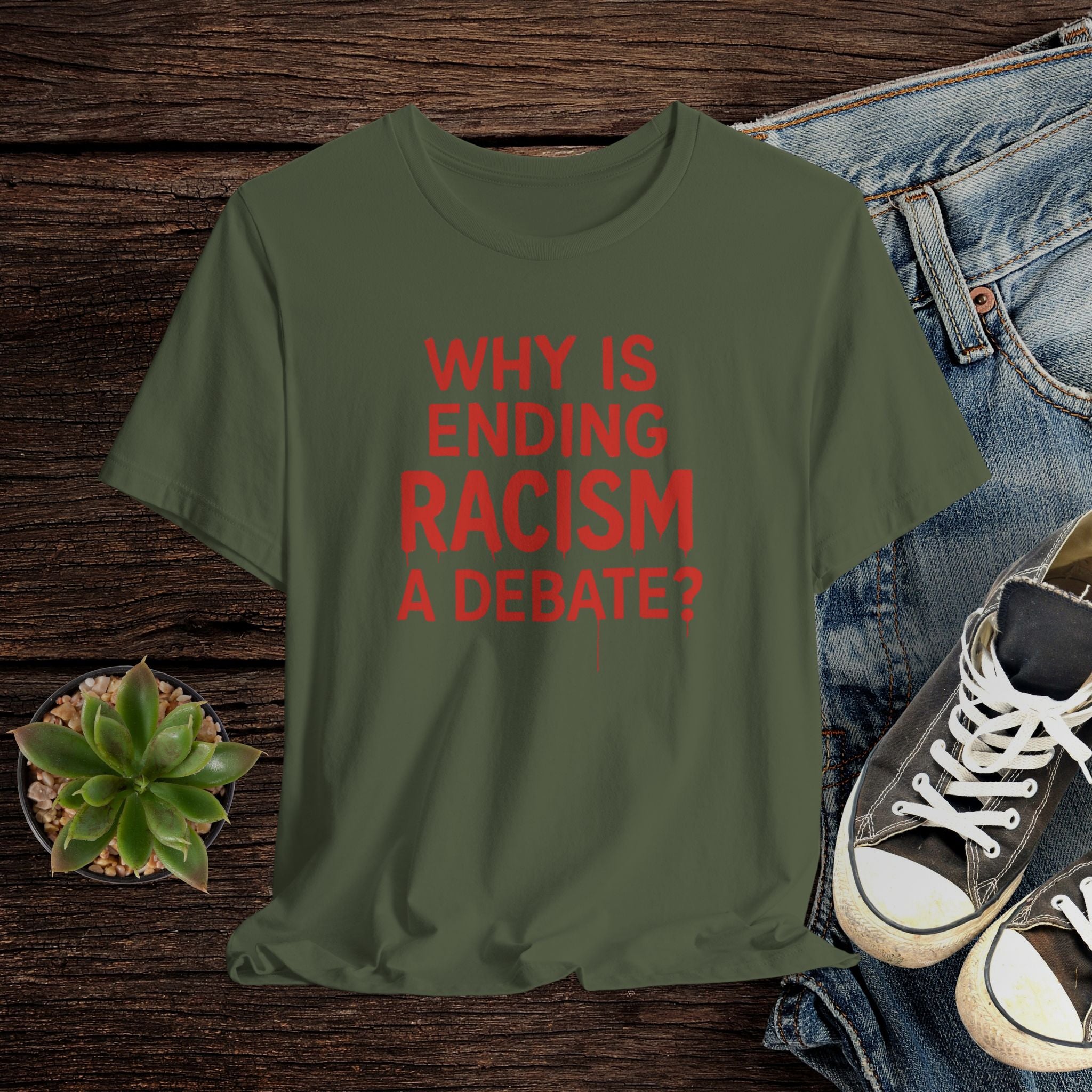 Green t-shirt with red text 'WHY IS ENDING RACISM A DEBATE?' on a wooden surface with jeans and sneakers.