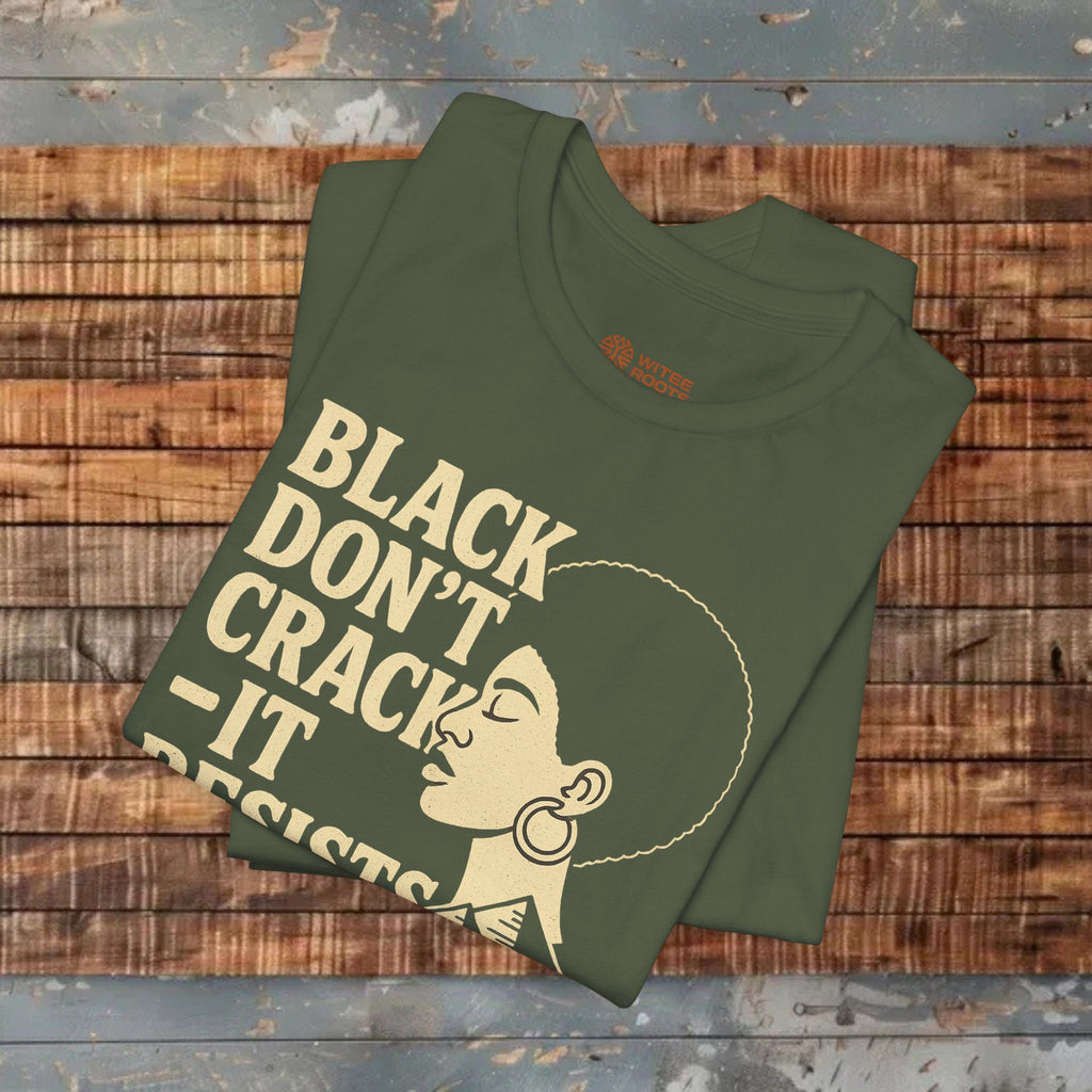 An olive green folded t-shirt with a cream graphic print of a woman's profile and the text 'BLACK DON'T CRACK - IT RESISTS' on a rustic wooden plank background.