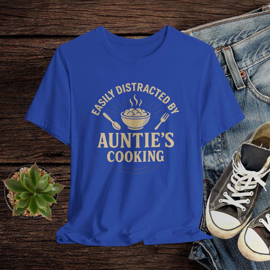 Blue t-shirt with 'Easily Distracted by Auntie's Cooking' text on a wooden surface with jeans and shoes.