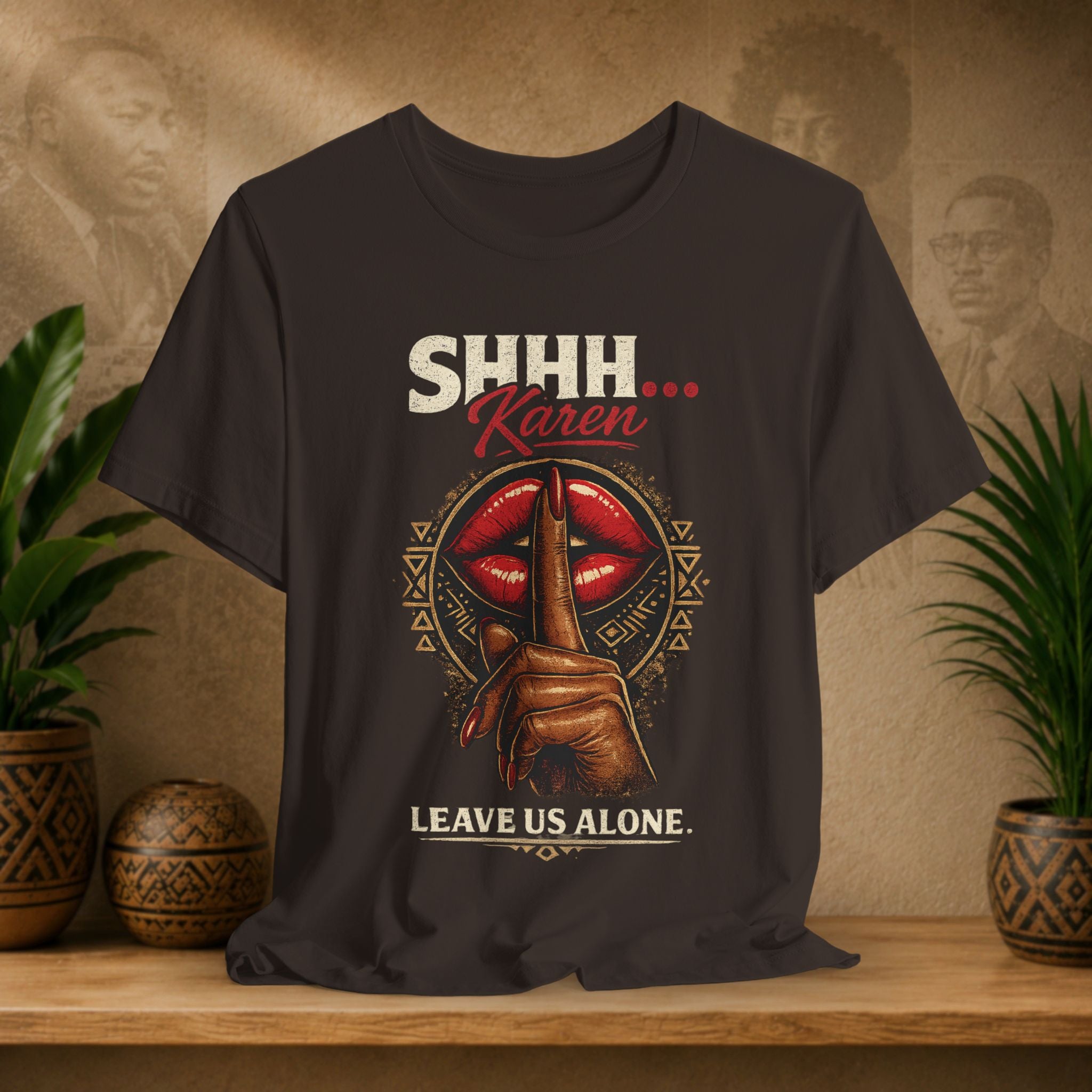 A brown graphic t-shirt displayed on a wooden shelf. The shirt features a graphic of a hand holding a finger vertically in front of red lips, with the text 'SHHH’.