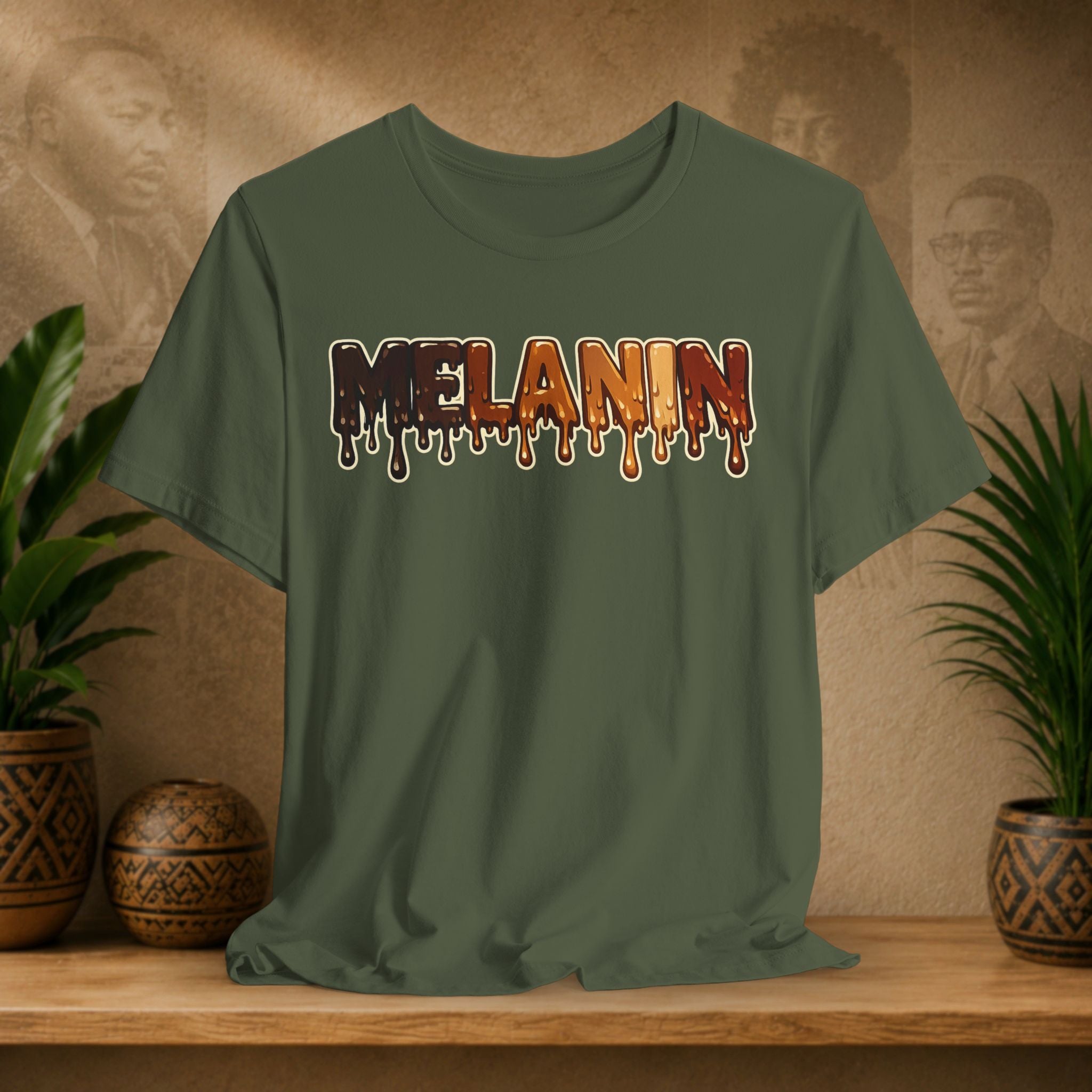 An olive green short-sleeve t-shirt with a dripping 'MELANIN' graphic in a brown-to-orange gradient, displayed on a wooden shelf with potted plants against a textured beige background.