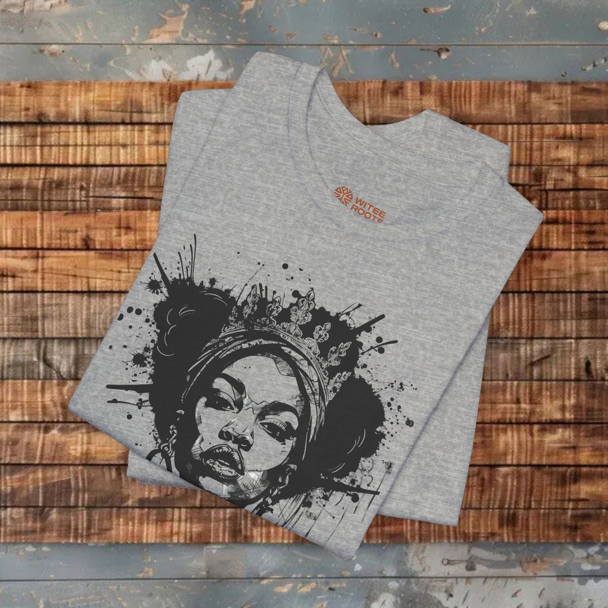 A folded heather gray t-shirt with a black graphic print of a woman's face surrounded by paint splatters, displayed on a rustic wooden plank surface.