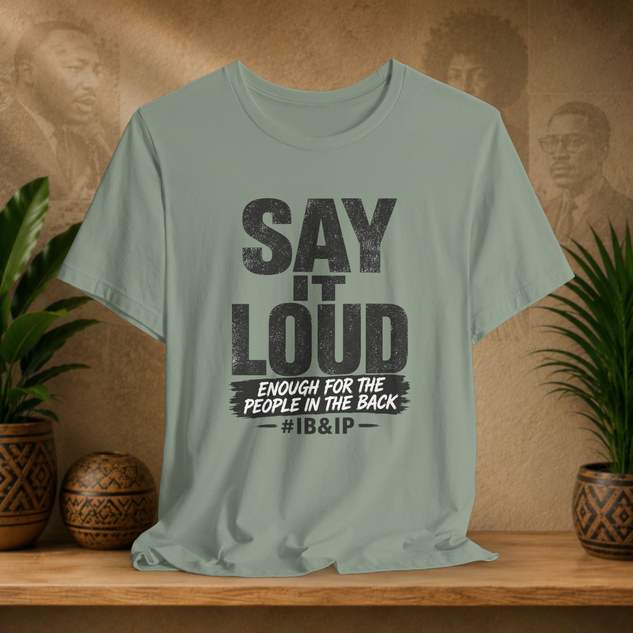 Green t-shirt with text 'Say it Loud Enough for the People in the Back #IB&IP' on a wooden surface with plants and a textured wall background.