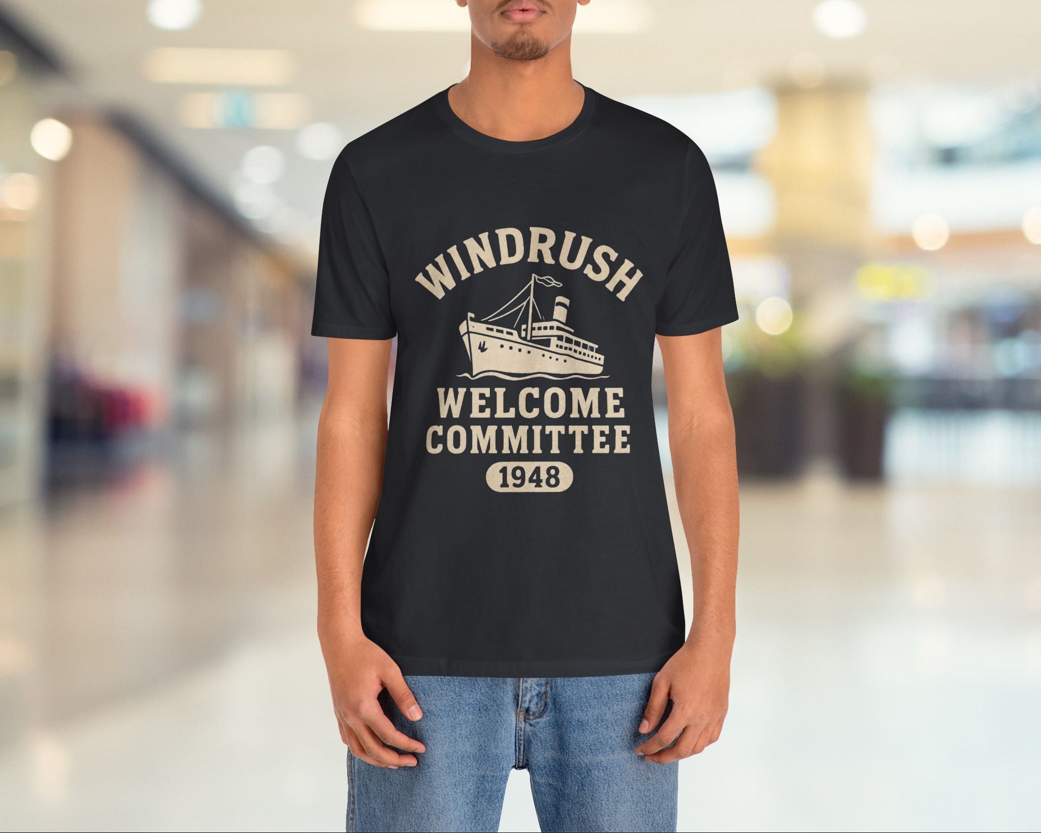 Person wearing a black t-shirt with 'Windrush Welcome Committee 1948' text and graphic in a blurred indoor setting.