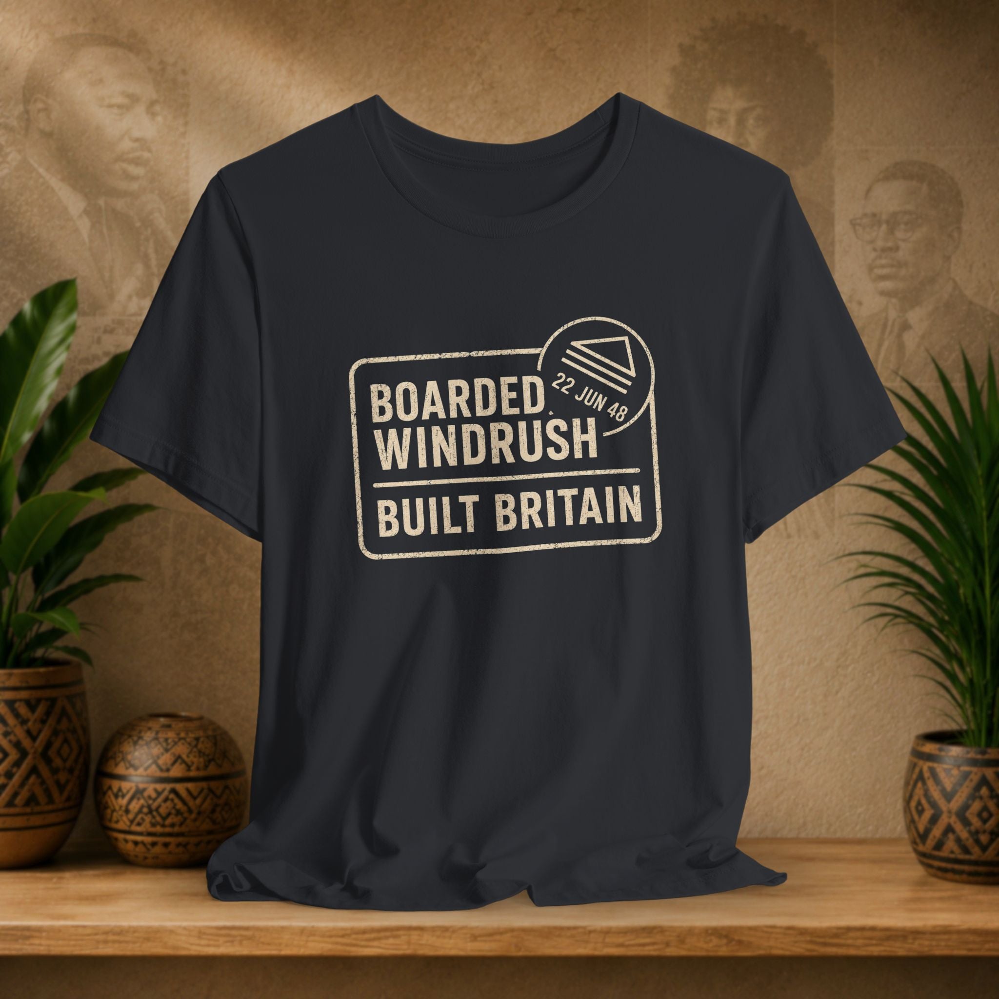 Black t-shirt with 'Boarded Windrush Built Britain' text on a wooden surface with plants and a textured wall background.