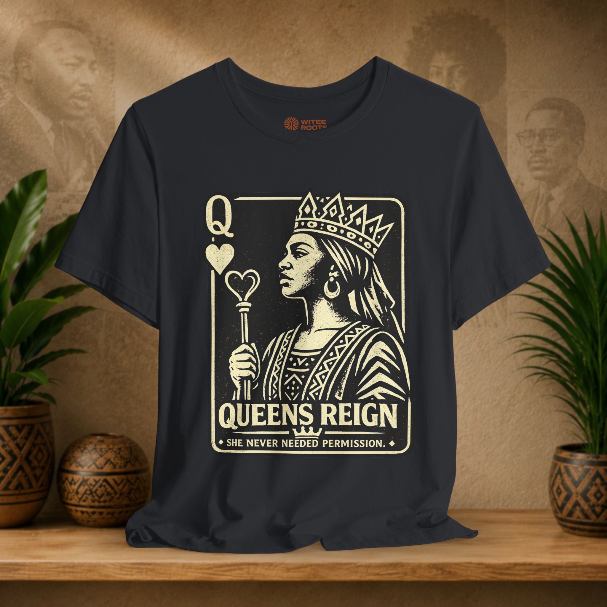 Black t-shirt with a graphic design of a queen holding a scepter and text on a wooden surface with plants and decorative items.