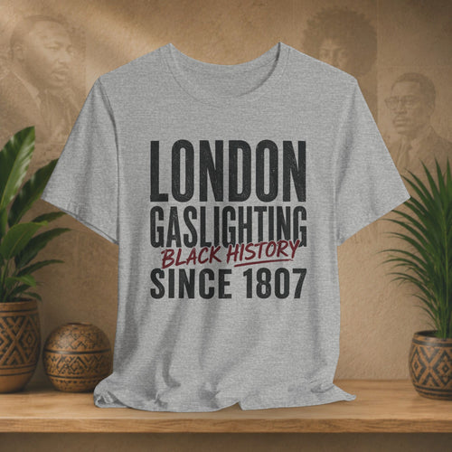 Gray t-shirt with 'London Gaslighting Black History Since 1807' text on a wooden surface with plants and blurred background