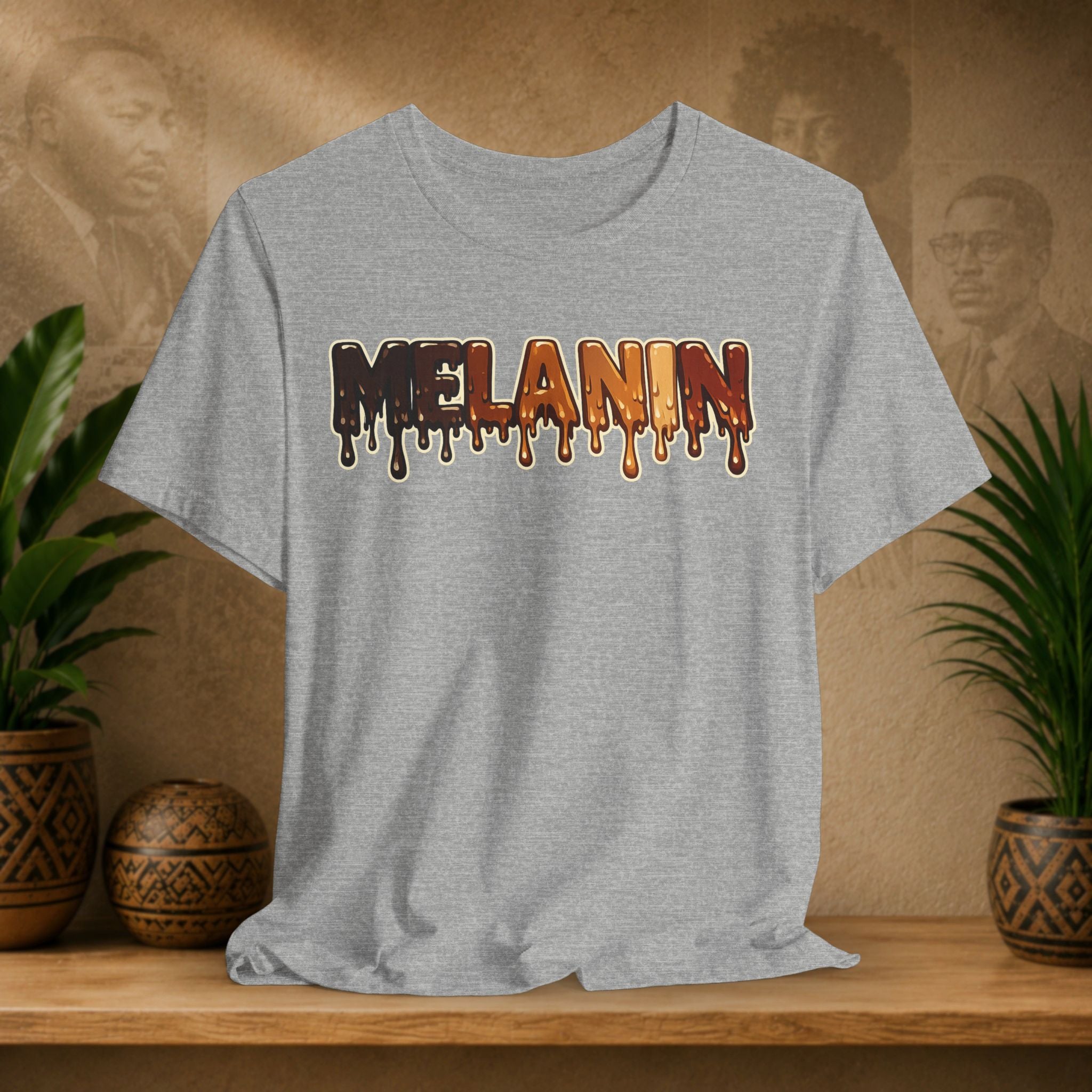 A light heather gray t-shirt with the word 'MELANIN' printed across the chest in a dripping paint style using shades of brown, orange, and red. The shirt is displayed on a wooden shelf in a styled setting with potted plants and a textured background.