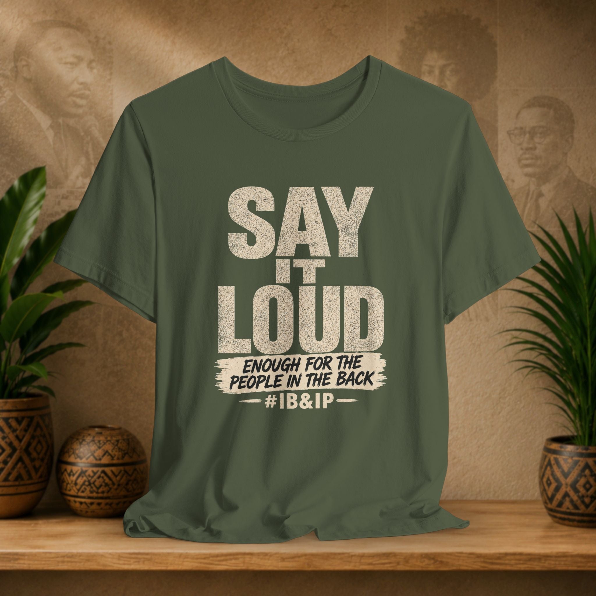Green t-shirt with text 'Say it Loud Enough for the People in the Back #IB&IP' on a wooden surface with plants and a textured wall background.