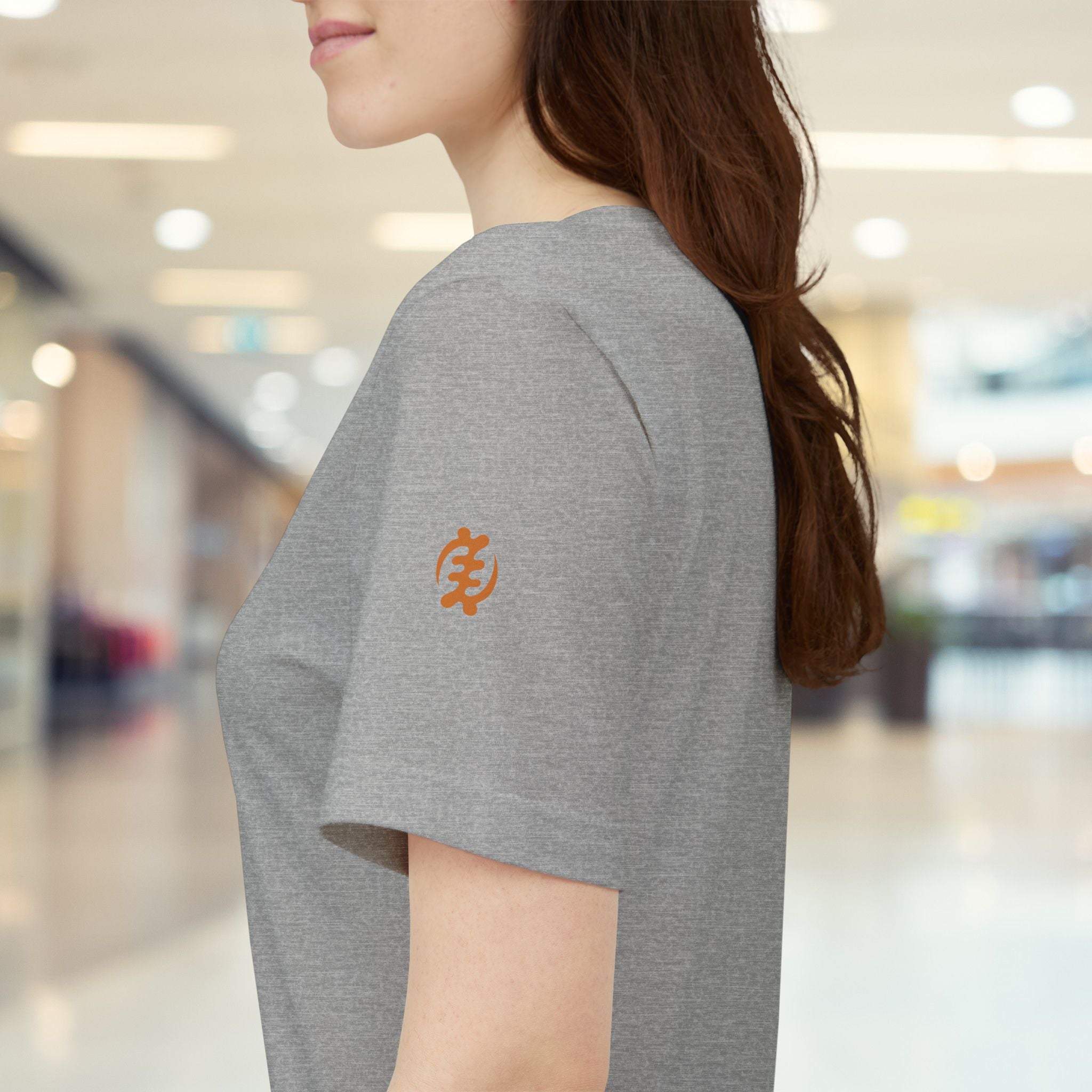 Person wearing a gray t-shirt with an orange logo in a blurred indoor setting