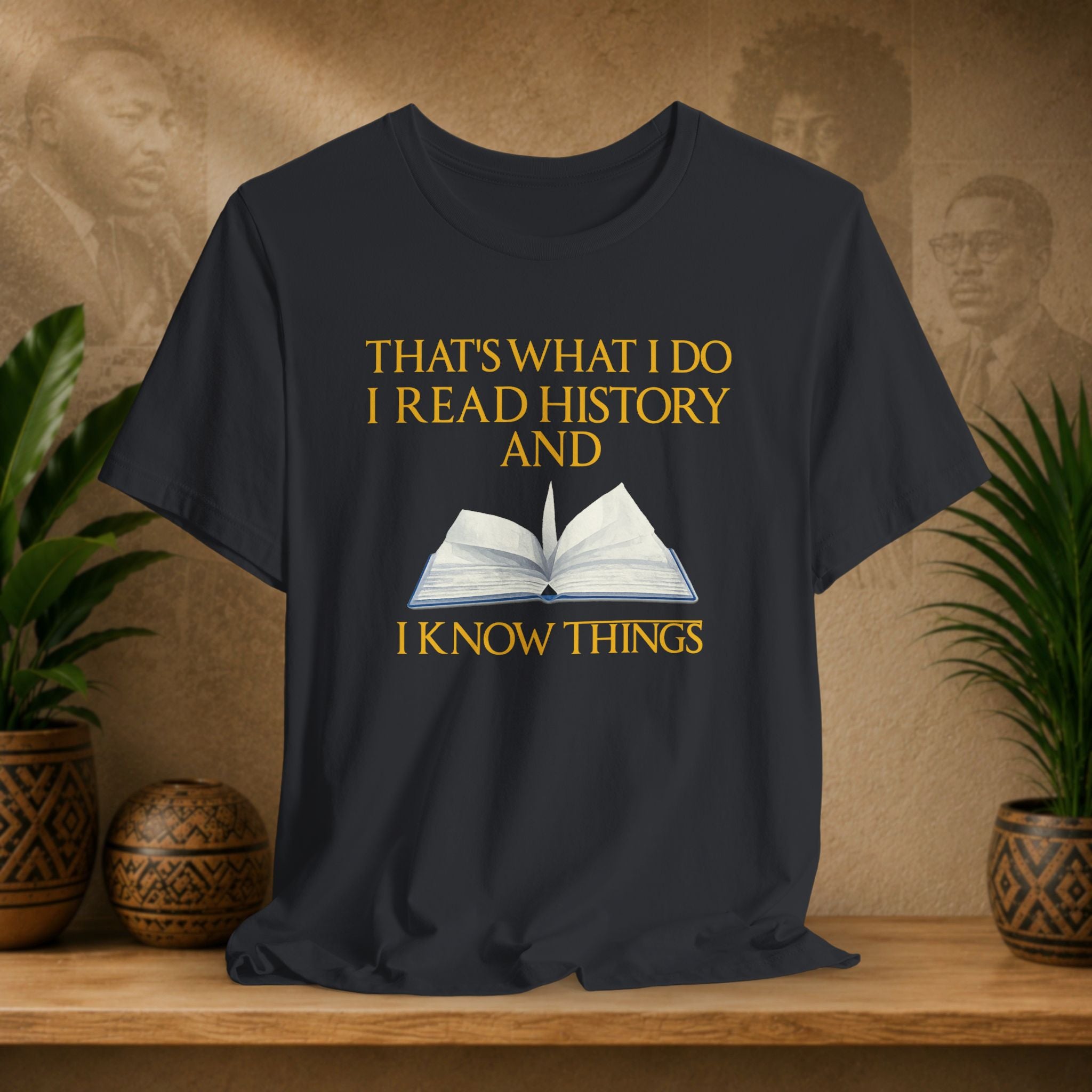 Black t-shirt with yellow text and book graphic on a wooden surface with plants and a textured wall background