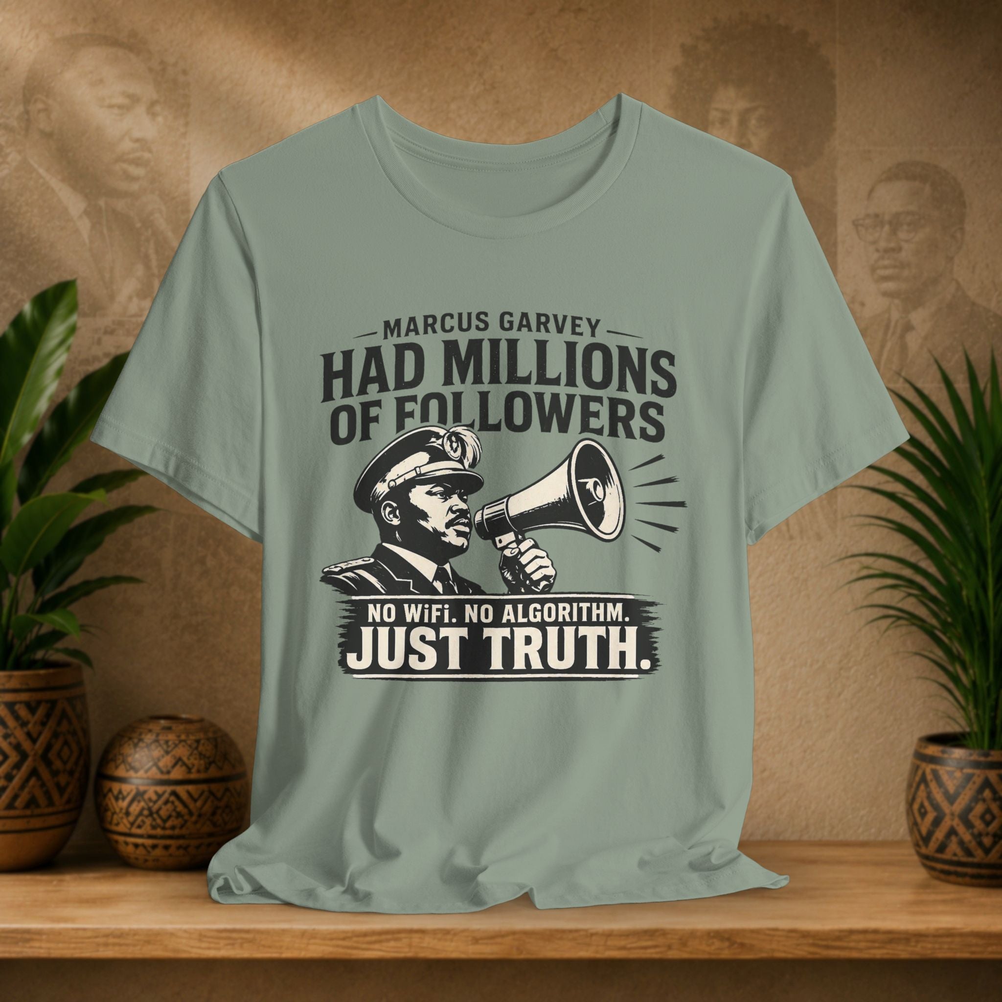 Sage green t-shirt with graphic and text on a wooden surface with plants and decorative items.