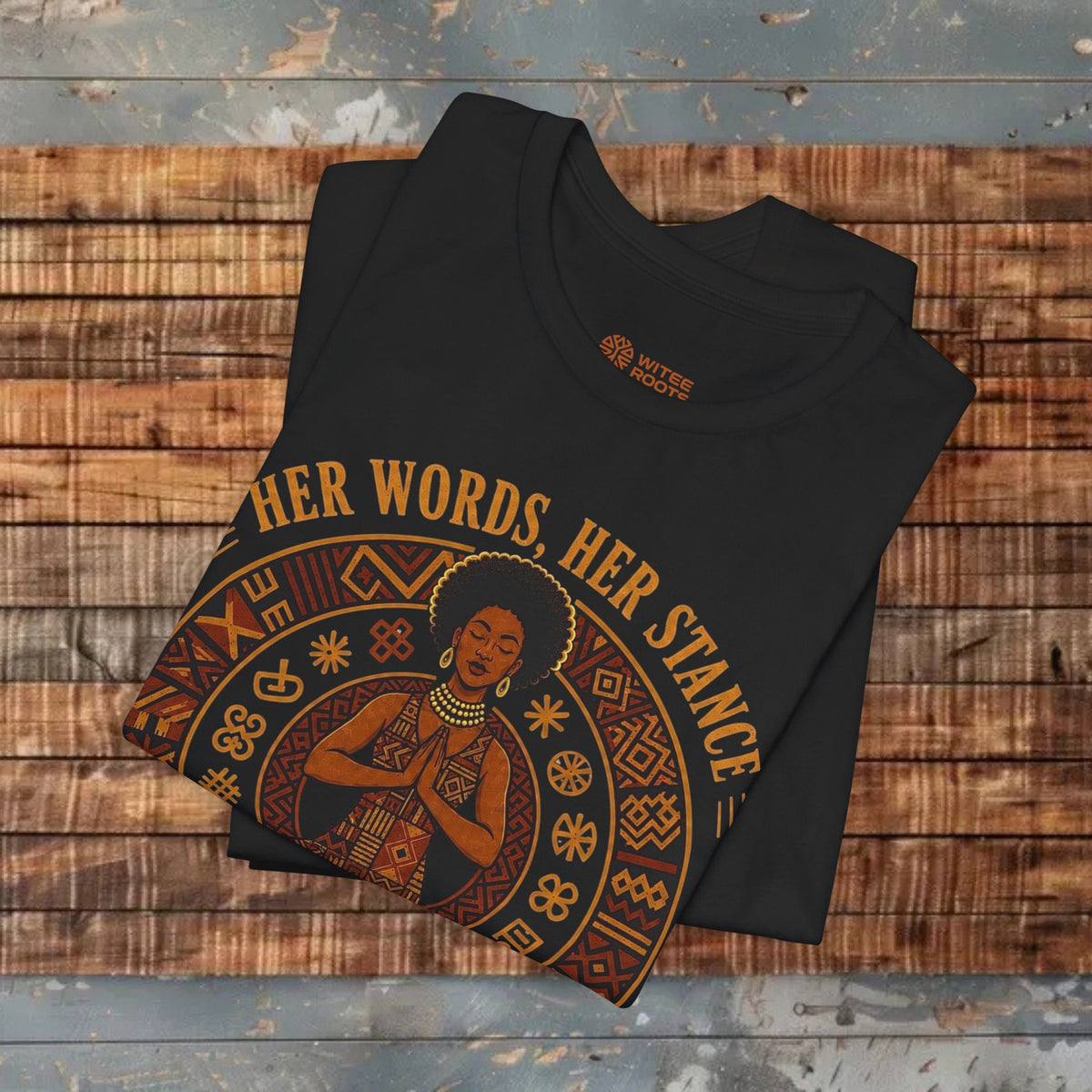 A folded black graphic T-shirt displayed on a rustic wooden plank surface. The shirt features a circular design with an illustration of a woman and the text 'OTHER WORDS HER STANCE' in a warm tan and orange tone.