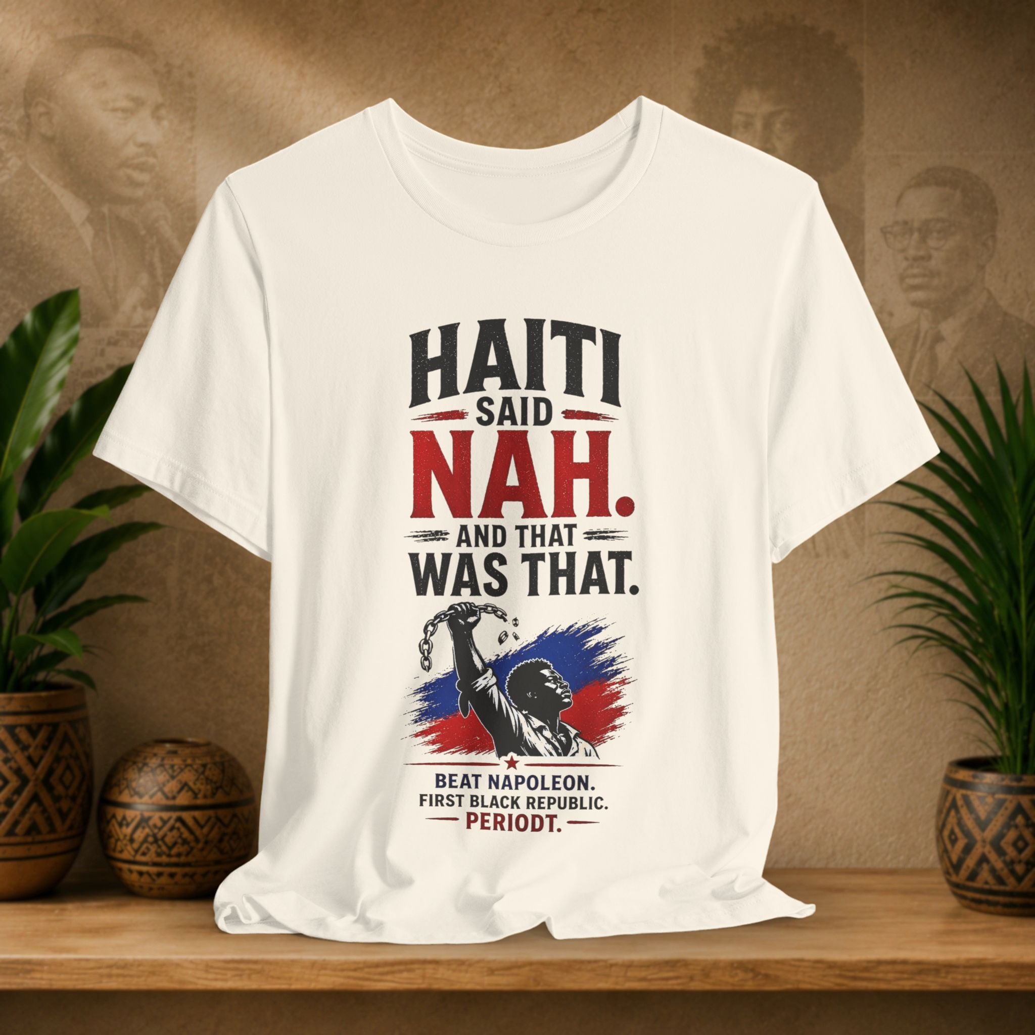 Beige t-shirt with Haiti-themed graphic and text on a wooden surface with plants and a textured wall background.