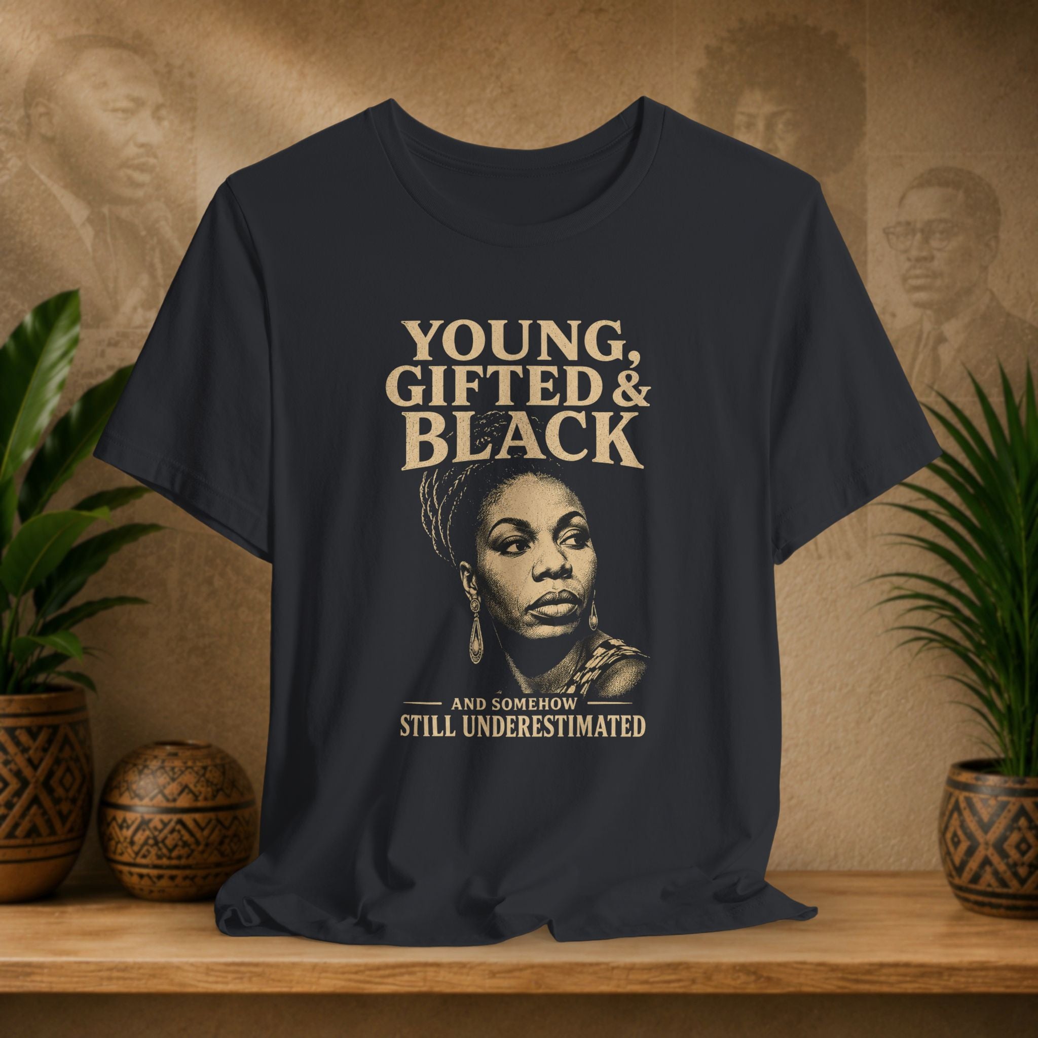 A black graphic t-shirt displayed on a wooden shelf. The shirt features a sepia-toned portrait of Audie Loney and the text 'YOUNG, GIFTED & BLACK' and 'AND SOMEHOW STILL UNDERESTIMATED' in a light beige font.