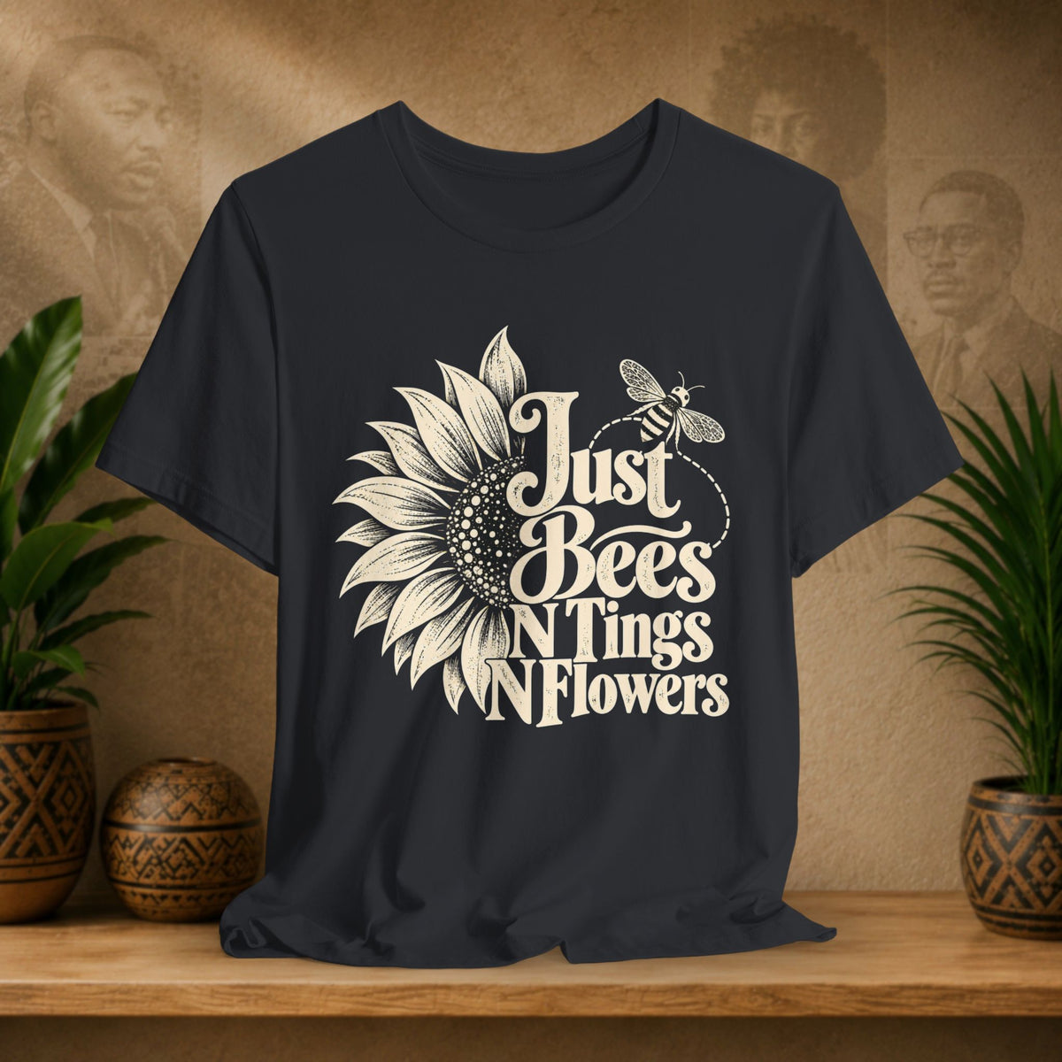 A black t-shirt with a cream-colored graphic of a sunflower, bee, and the text 'Just Bees N Tings N Flowers', displayed on a wooden shelf with potted plants and a textured background.