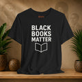 Black t-shirt with 'BLACK BOOKS MATTER' text on a wooden surface with plants and a textured wall background.