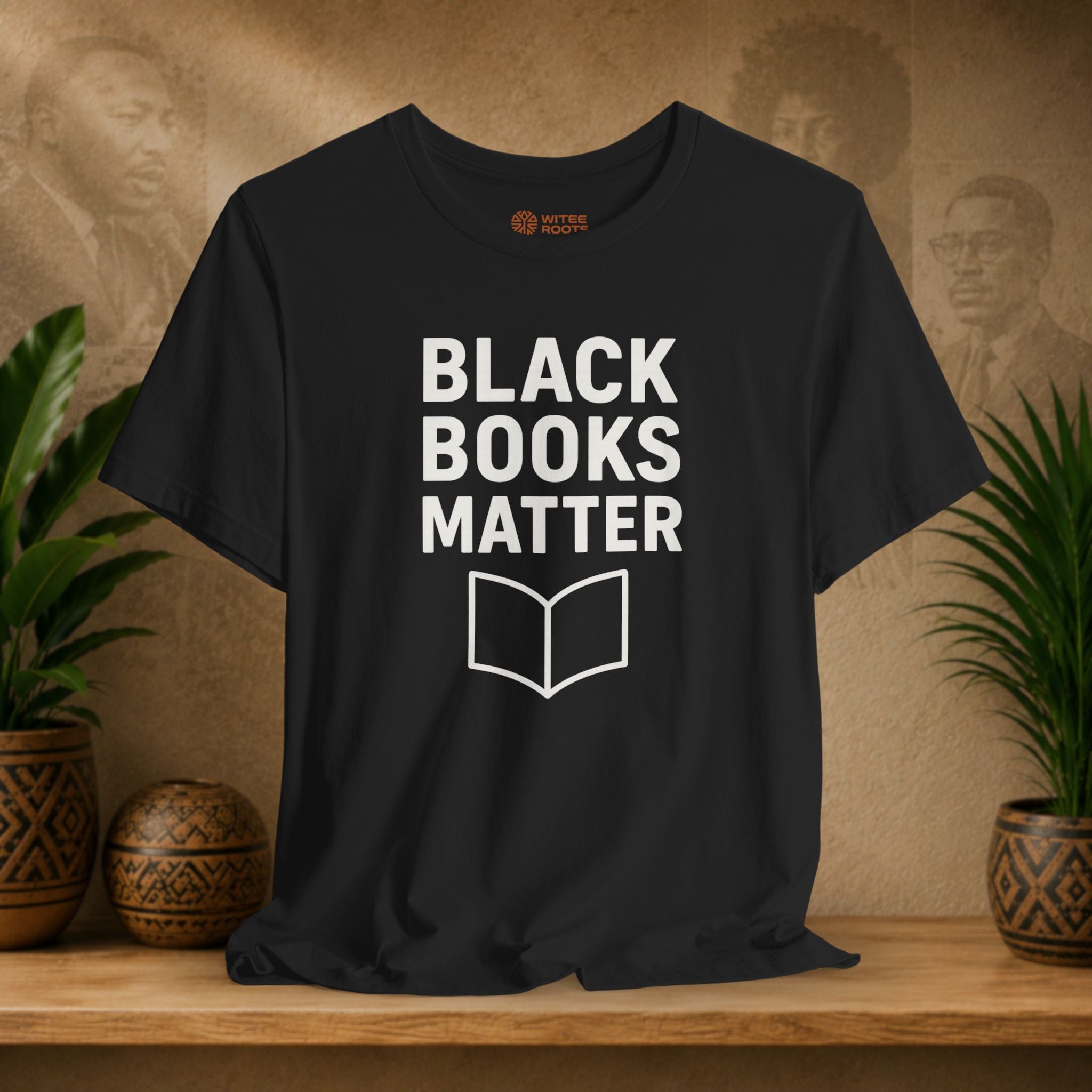 Black t-shirt with 'BLACK BOOKS MATTER' text on a wooden surface with plants and a textured wall background.