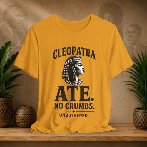 Yellow t-shirt with Cleopatra graphic and text on a wooden surface with plants and decorative items.