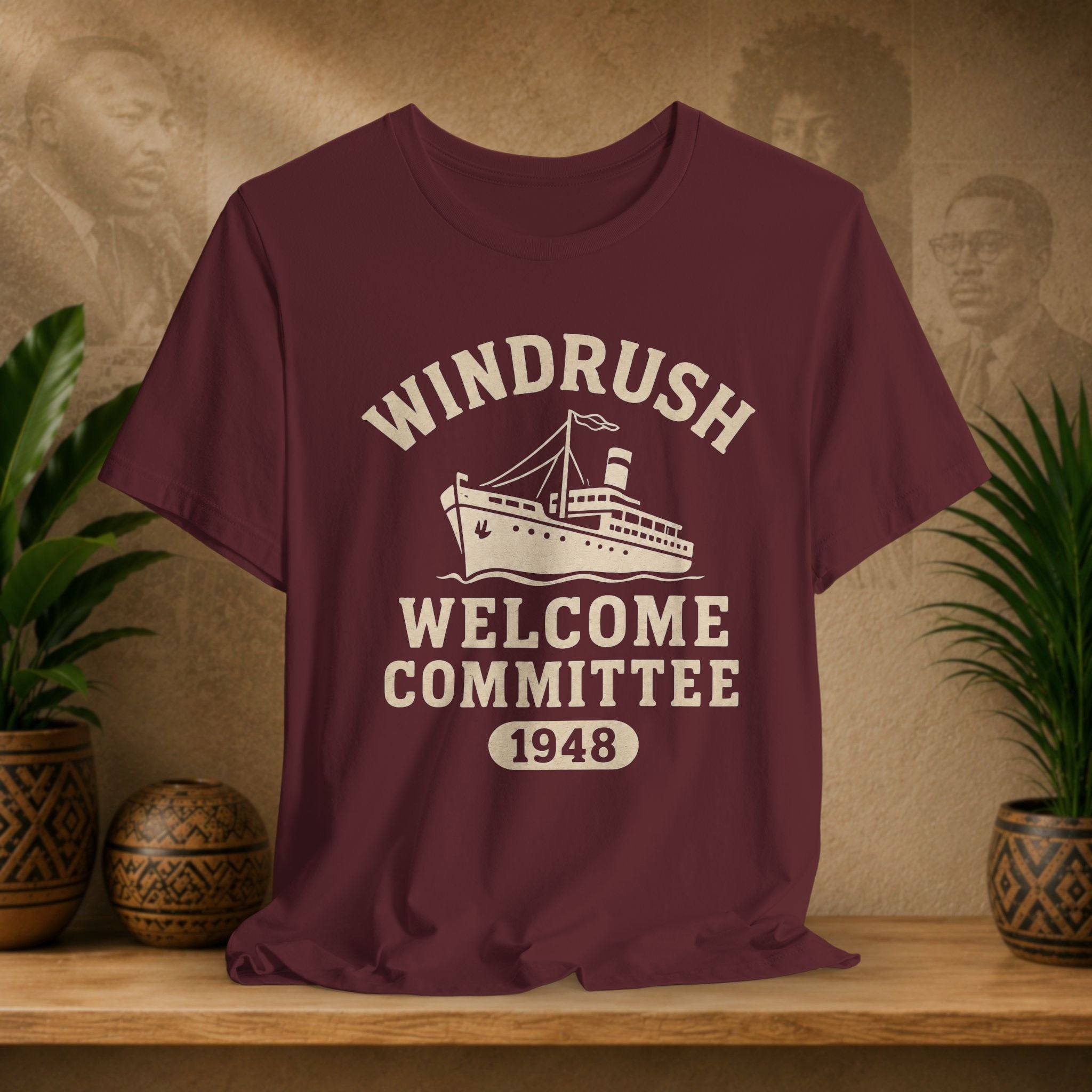 Maroon t-shirt with 'Windrush Welcome Committee 1948' design on a wooden surface with plants and a decorative wall.